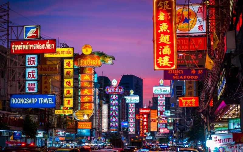 Scène animée de Chinatown à Bangkok, illuminée par des enseignes lumineuses colorées en caractères chinois et thaïlandais au crépuscule